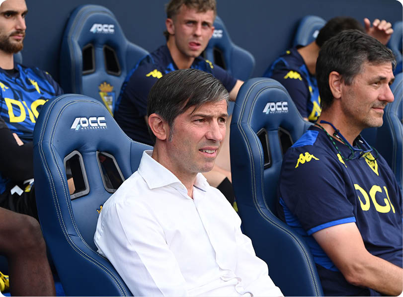 Several people wearing matching sports uniforms sit in a row of blue stadium seats, with one person in a white shirt seated prominently in the foreground. Descripci n generada con IA