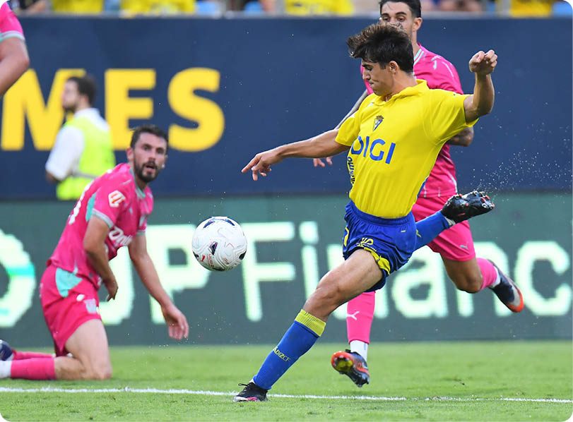 A person in a yellow and blue sports uniform is kicking a soccer ball while two people in pink uniforms are nearby on a grassy field during a game. Descripci n generada con IA