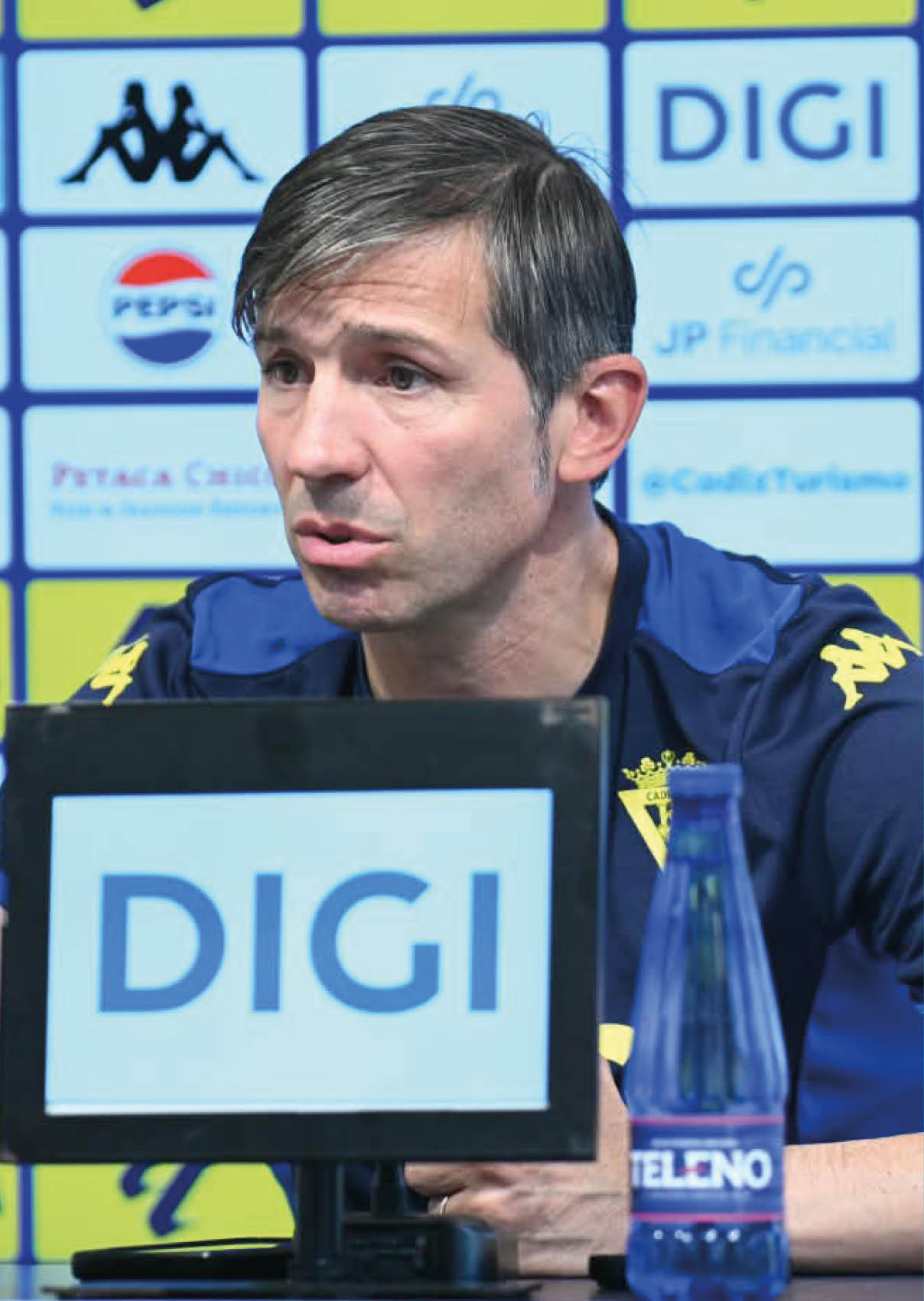 Una persona con camiseta deportiva azul con detalles amarillos est sentada en una mesa frente a un micr fono con un cartel que dice DIGI y varios logotipos de patrocinadores en el fondo.  Descripci n generada con IA