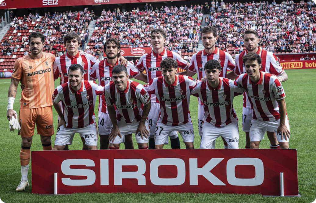 Equipo de f tbol con uniforme a rayas rojas y blancas posando en el campo antes de un partido con un cartel rojo que dice SIROKO frente a ellos y p blico en las gradas al fondo. Descripci n generada con IA