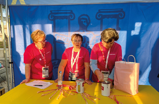 Tres personas con camisetas rojas est n detr s de una mesa amarilla con objetos como botellas, latas y una bolsa de papel en un entorno con fondo azul. Descripci n generada con IA