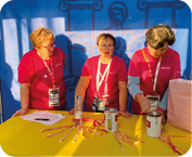 Tres personas adultas con camisetas rojas y gafetes est n detr s de una mesa amarilla con cintas y latas para donaciones en un evento. Descripci n generada con IA