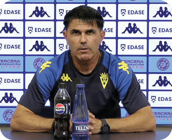 Una persona con camiseta deportiva azul y negra con logos amarillos est sentada frente a un micr fono en una mesa con botellas de agua y refresco, con un fondo de paneles publicitarios deportivos.  Descripci n generada con IA