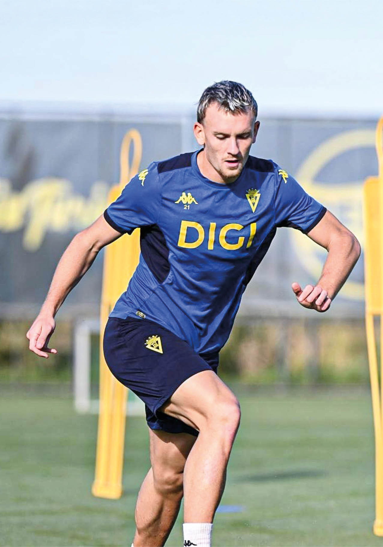 Persona con camiseta y pantalones deportivos azules entrenando en un campo de f tbol con conos amarillos de fondo. Descripci n generada con IA