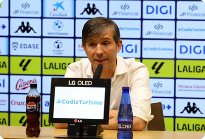 A person with short hair wearing a white shirt is speaking into a microphone at a press conference table with a digital screen displaying “@CadizTurismo" and bottles of Pepsi and water in front. Descripci n generada con IA