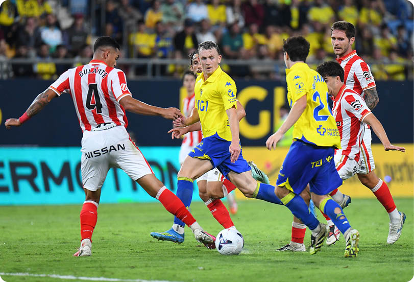 People wearing yellow and blue uniforms and others in red and white striped uniforms are actively competing for a soccer ball on a grass field during a match. Descripci n generada con IA