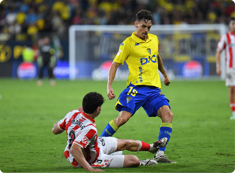A person in a yellow and blue sports uniform is dribbling a soccer ball while another person in a red and white striped uniform is sliding on the grass attempting to tackle the ball. Descripci n generada con IA