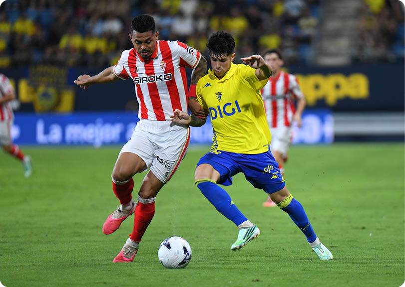 Two adults in sports uniforms compete for a soccer ball on a grassy field during a match with a blurred crowd in the background. Descripci n generada con IA