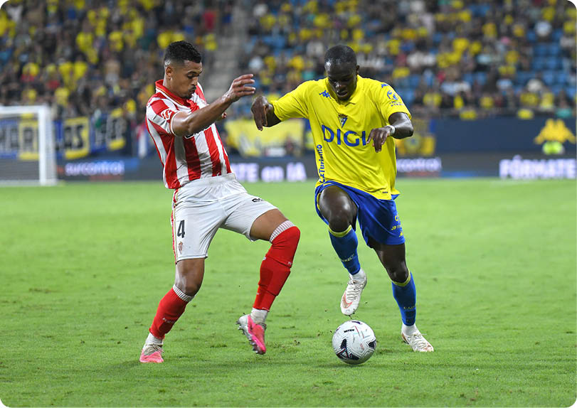 Two people in sports uniforms compete for a soccer ball on a grass field with a crowd in the background. Descripci n generada con IA