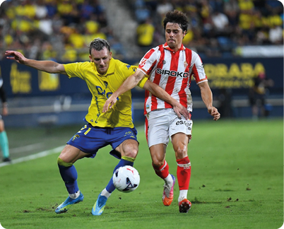 Two people in sports uniforms compete for a soccer ball on a grass field during a match with spectators in the background. Descripci n generada con IA