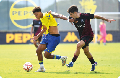 Two people in sports uniforms are competing for a soccer ball on a grassy field with one person pulling the other's shirt. Descripci n generada con IA