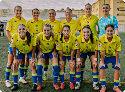 A group of eleven people wearing yellow and blue soccer uniforms are posing together on a soccer field with buildings in the background. Descripci n generada con IA