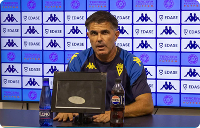 A person wearing a sports jersey is sitting at a table with a microphone and two bottles in front of a backdrop covered with logos. Descripci n generada con IA