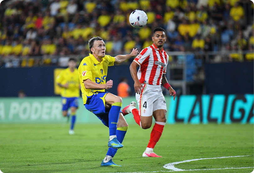 Two people in sports uniforms are actively playing soccer on a field with one person in a yellow and blue outfit kicking the ball while the other in a red and white striped outfit runs nearby. Descripci n generada con IA