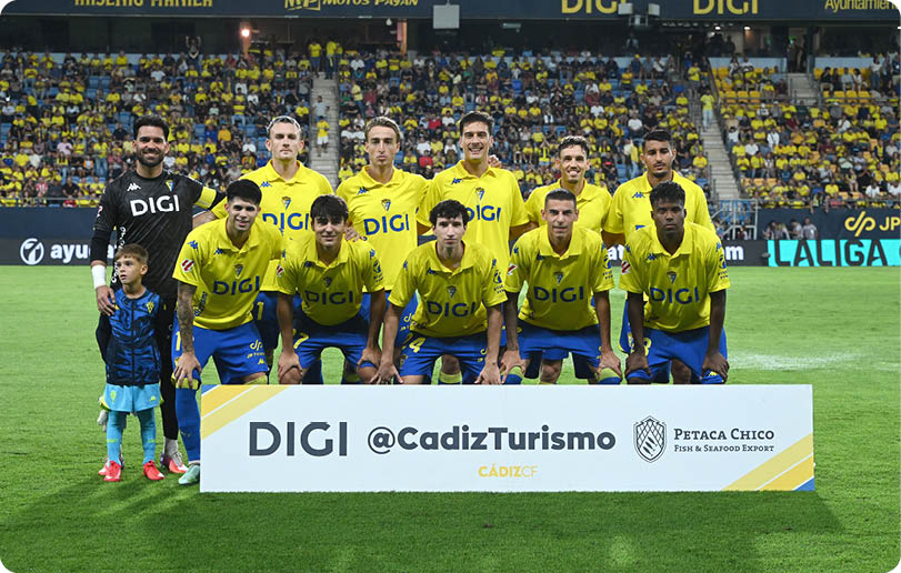 A sports team wearing yellow and blue uniforms poses on a soccer field behind a sign with sponsor logos and a crowd in the stadium stands behind them. Descripci n generada con IA
