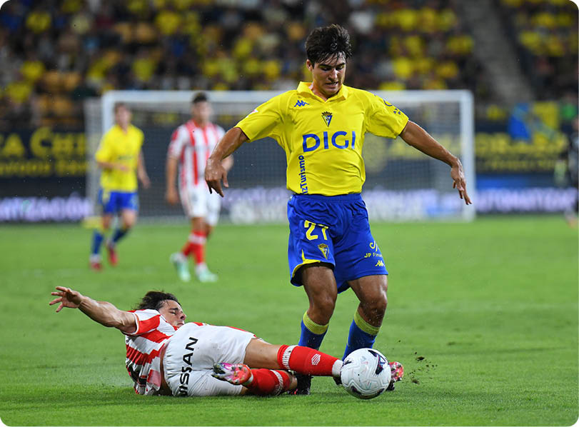 A person in a yellow and blue soccer uniform is controlling the ball while another person in a red and white striped uniform is sliding on the grass attempting to tackle the ball on a soccer field. Descripci n generada con IA