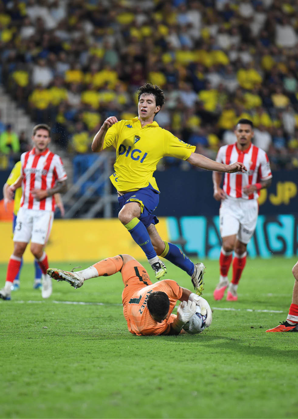 A person in a yellow and blue soccer uniform jumps over a goalkeeper in an orange outfit who is on the ground holding a soccer ball while other players in red and white striped uniforms watch in the background. Descripci n generada con IA