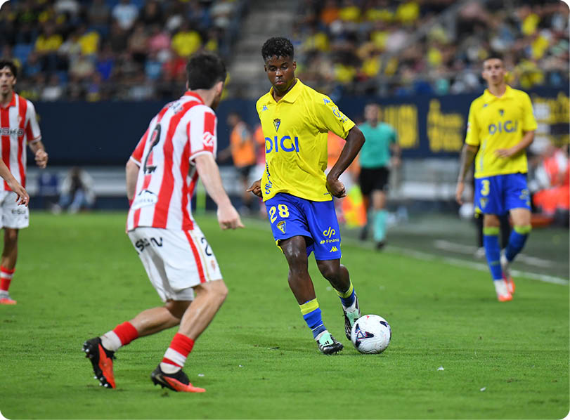 People wearing yellow and blue sports uniforms and red and white striped uniforms are playing soccer on a grass field in a stadium with spectators in the background. Descripci n generada con IA