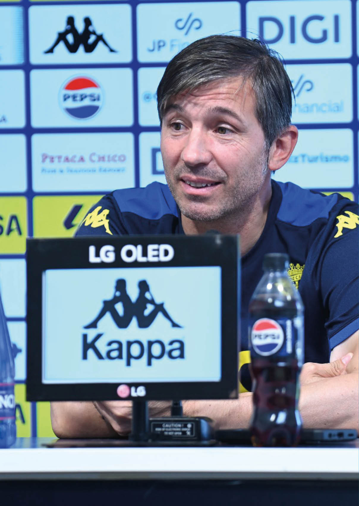 Una persona con camiseta deportiva azul con logos amarillos est sentada frente a un micr fono y una botella de refresco en una conferencia de prensa.  Descripci n generada con IA