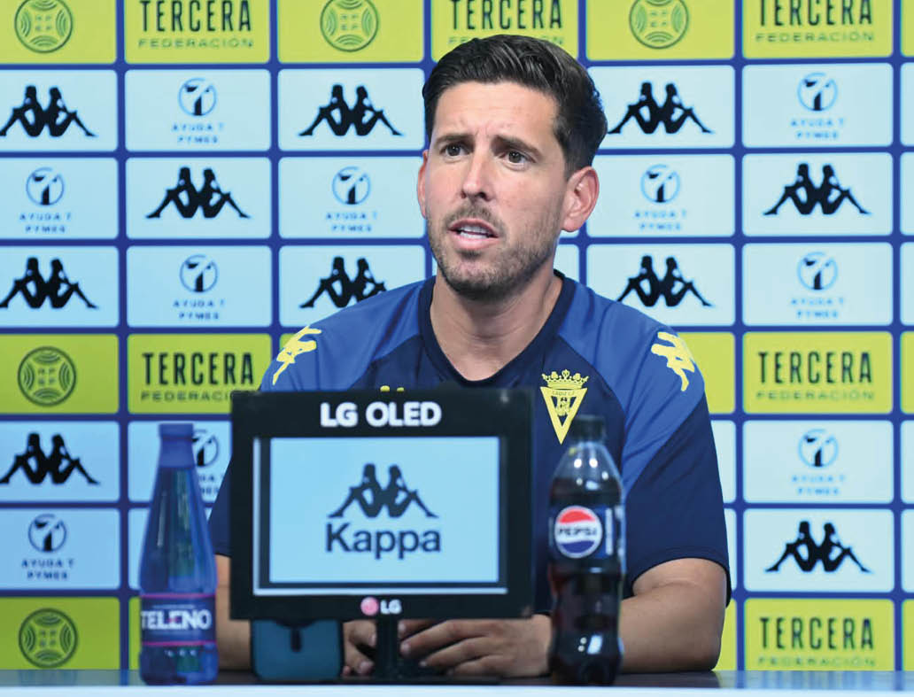 Una persona con camiseta deportiva azul est sentada en una mesa frente a un micr fono y una botella de refresco en una conferencia de prensa con un fondo de logotipos deportivos.  Descripci n generada con IA
