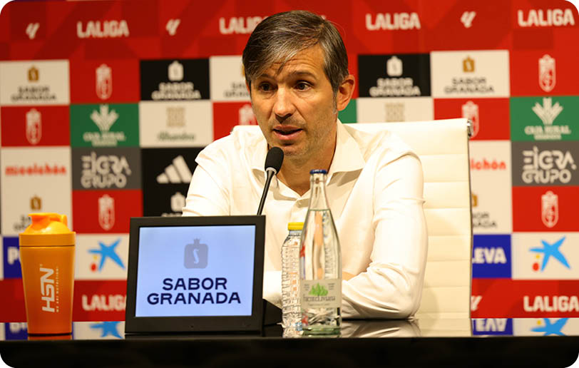 A person in a white shirt is speaking into a microphone at a press conference with a backdrop featuring various logos and a screen displaying the text “SABOR GRANADA." Descripci n generada con IA