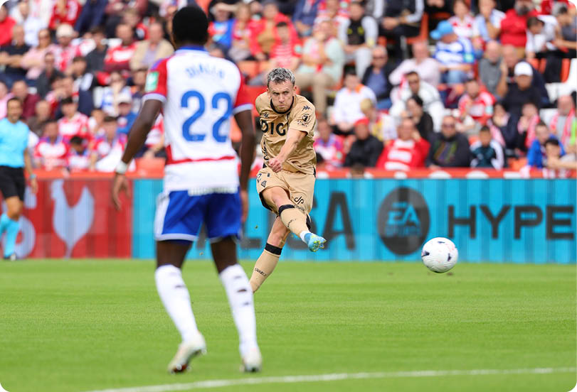 A person in a gold sports uniform is kicking a soccer ball while another person in a white and blue uniform watches on a grassy field with a crowd in the background. Descripci n generada con IA