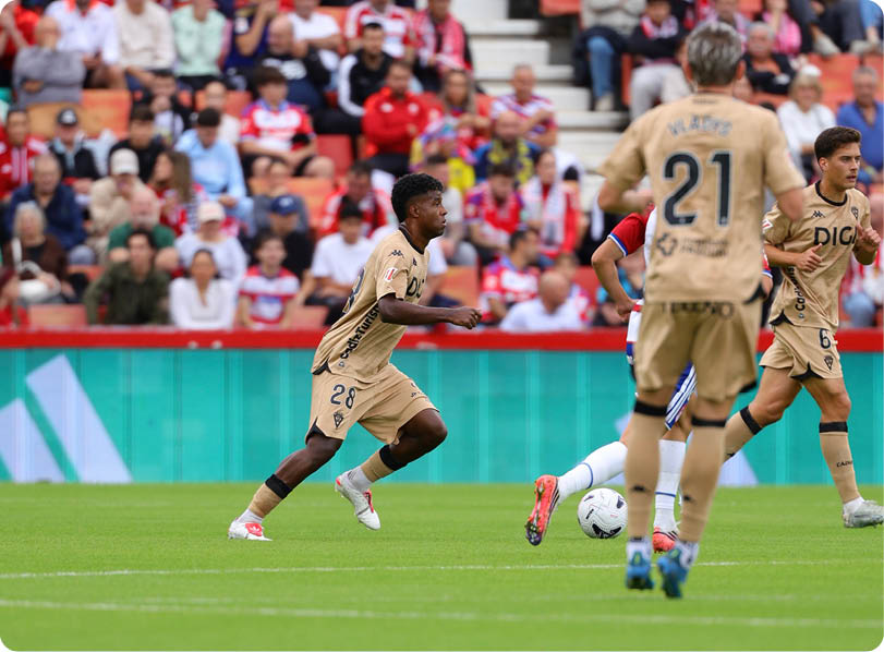 People wearing beige sports uniforms are actively playing soccer on a green field with a crowd of spectators in the background. Descripci n generada con IA