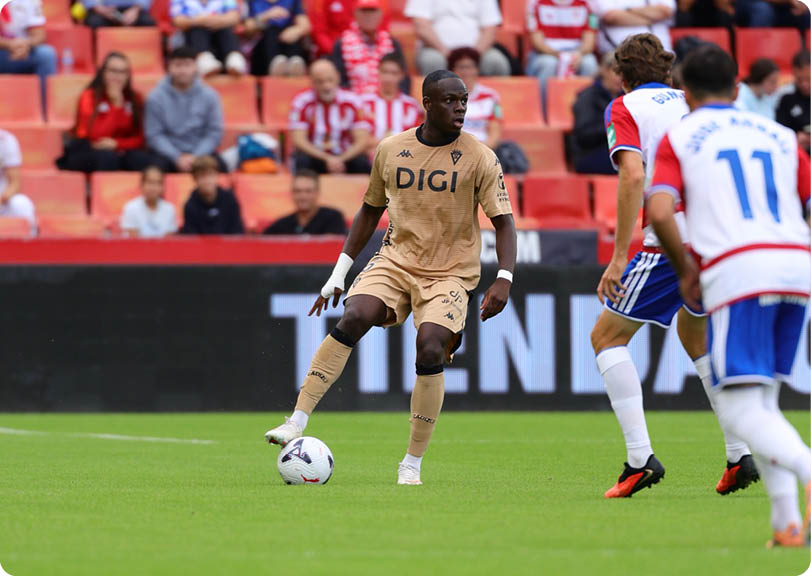 A person in a beige soccer uniform is controlling a soccer ball on a field while two people in white and blue uniforms approach. Descripci n generada con IA