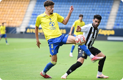 Two people in sports uniforms compete for a soccer ball on a grassy field inside a stadium with empty blue and yellow seats. Descripci n generada con IA