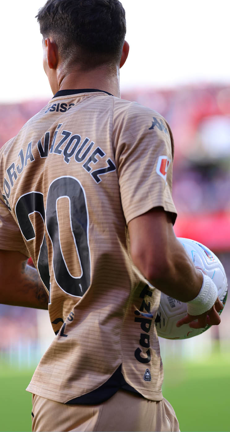 A person wearing a brown sports jersey with a wristband is holding a white soccer ball. Descripci n generada con IA