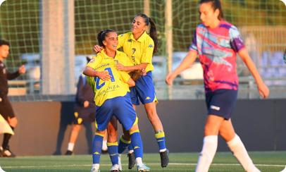 Two people in yellow sports uniforms are celebrating on a soccer field while another person in a colorful jersey walks nearby. Descripci n generada con IA