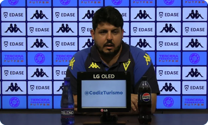 A person wearing a blue and black sports jacket is sitting at a press conference table with a microphone and two bottles, one labeled Pepsi, in front of a backdrop featuring logos and text related to Cadiz Turismo and Tercera Federaci n FutFem. Descripci n generada con IA