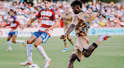 Two people in sports uniforms are actively playing soccer on a grassy field with one person about to kick the ball. Descripci n generada con IA