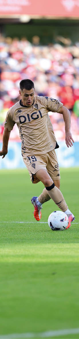A person wearing a beige soccer uniform with the number 19 is actively kicking a soccer ball on a green field with a blurred crowd in the background. Descripci n generada con IA