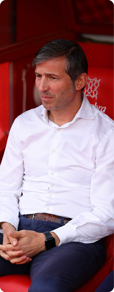A person with short dark hair wearing a white button-up shirt and dark pants is sitting on a red bench with hands clasped and a thoughtful expression. Descripci n generada con IA