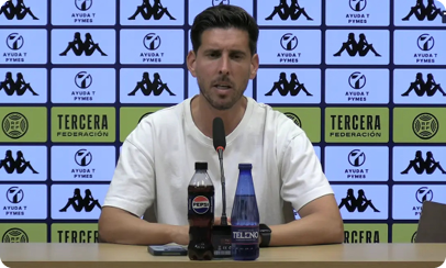 A person wearing a white t-shirt is sitting at a table with a microphone, a bottle of Pepsi, and a blue bottle in front of a backdrop featuring logos and text. Descripci n generada con IA