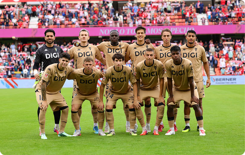 A group of eleven people wearing matching beige sports uniforms with “DIGI" on the front are posing together on a green soccer field with a crowd in the background. Descripci n generada con IA