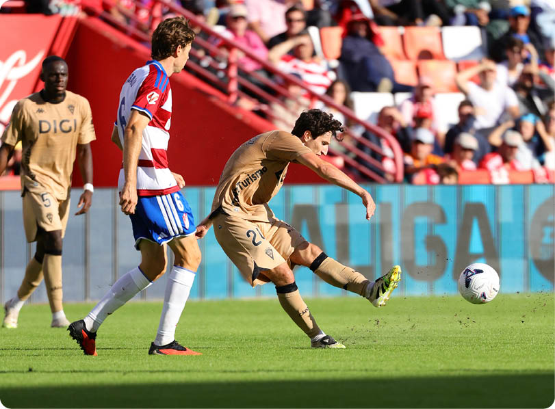 A person in a gold sports uniform is kicking a soccer ball while another person in a red, white, and blue striped uniform watches on a sunny soccer field with spectators in the background. Descripci n generada con IA
