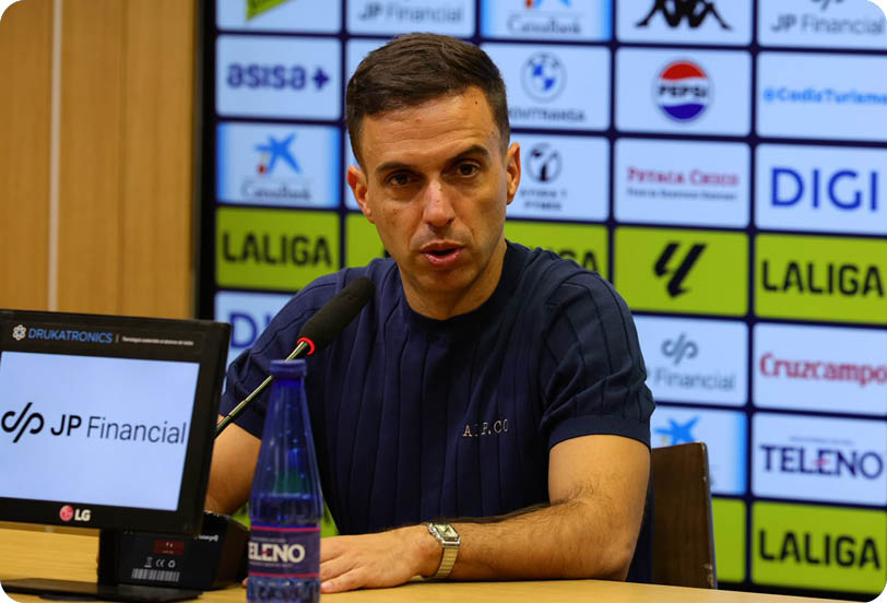 A person with short dark hair wearing a dark blue shirt is speaking into a microphone at a press conference with a backdrop featuring various sponsor logos. Descripci n generada con IA
