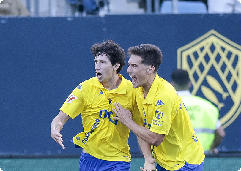Two adults wearing yellow sports jerseys are celebrating energetically on a sports field. Descripci n generada con IA