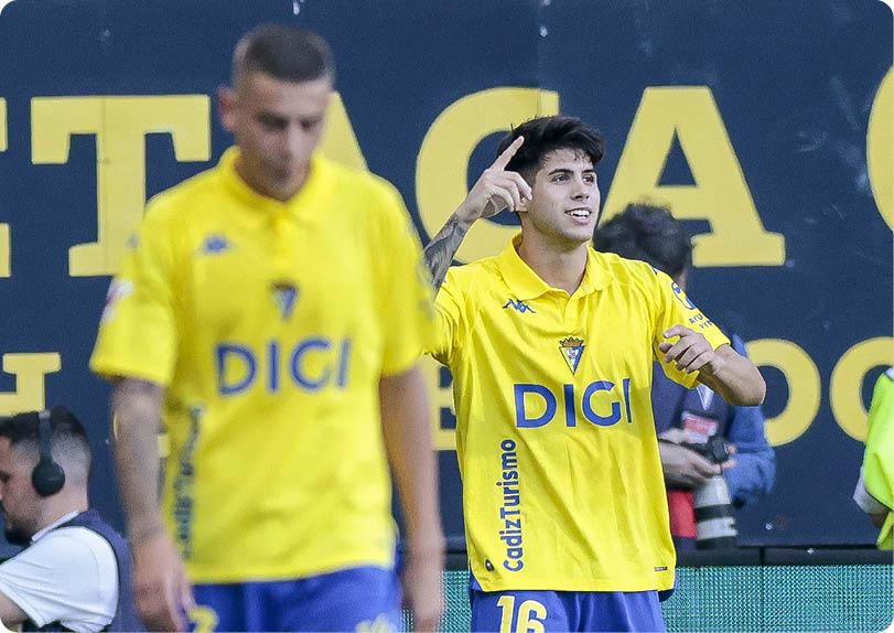 Two adults wearing yellow sports jerseys with blue shorts are on a field, with one person gesturing and the other walking with a blurred background of a sign and other people. Descripci n generada con IA