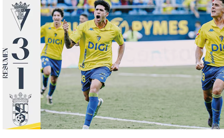People in yellow sports uniforms are celebrating on a soccer field with a scoreboard showing a 3-1 score. Descripci n generada con IA