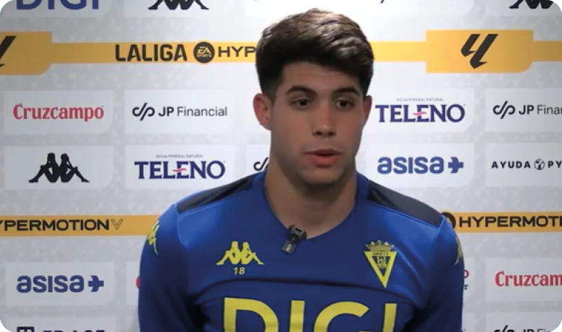 A person wearing a blue sports jersey with yellow logos and a microphone clipped to the collar is standing in front of a backdrop with various sponsor logos. Descripci n generada con IA
