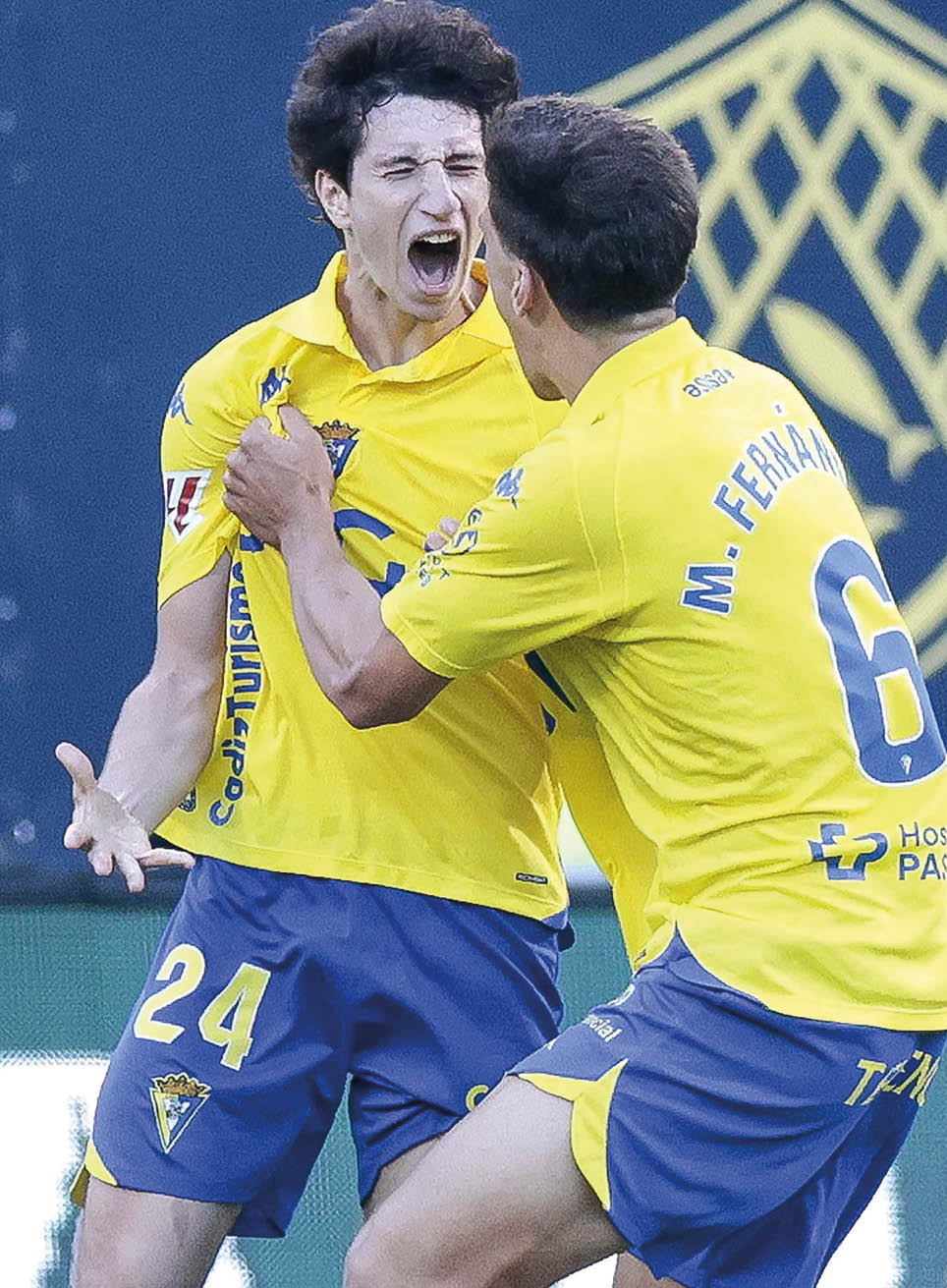 Two people wearing yellow and blue sports uniforms are celebrating energetically on a soccer field. Descripci n generada con IA