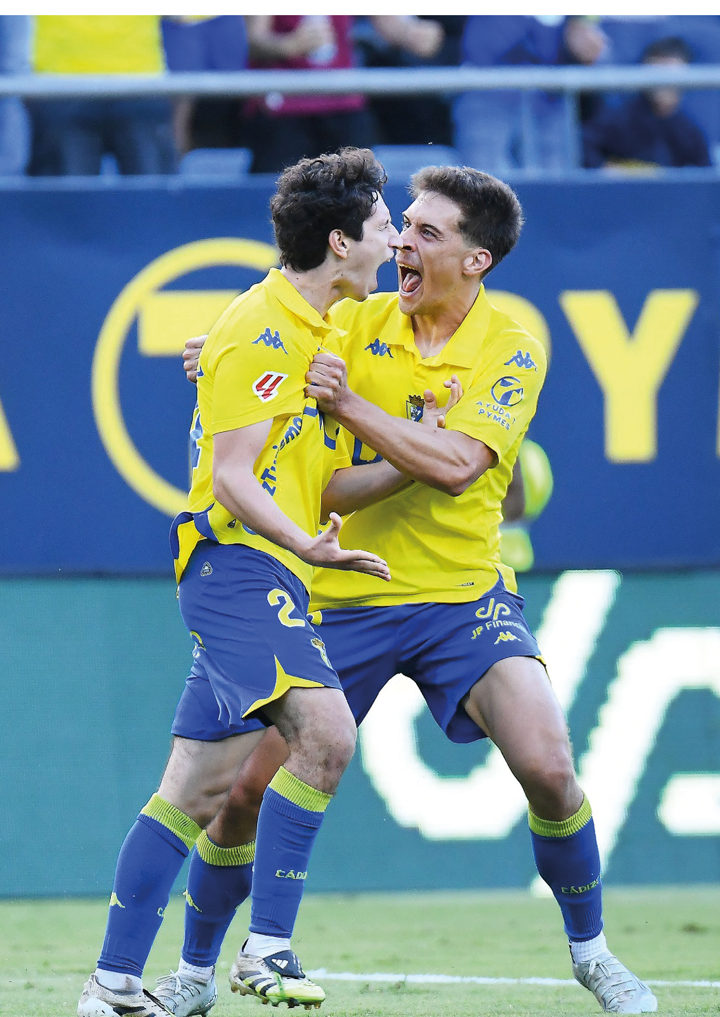 Two adults wearing yellow sports jerseys and blue shorts are celebrating energetically on a soccer field. Descripci n generada con IA