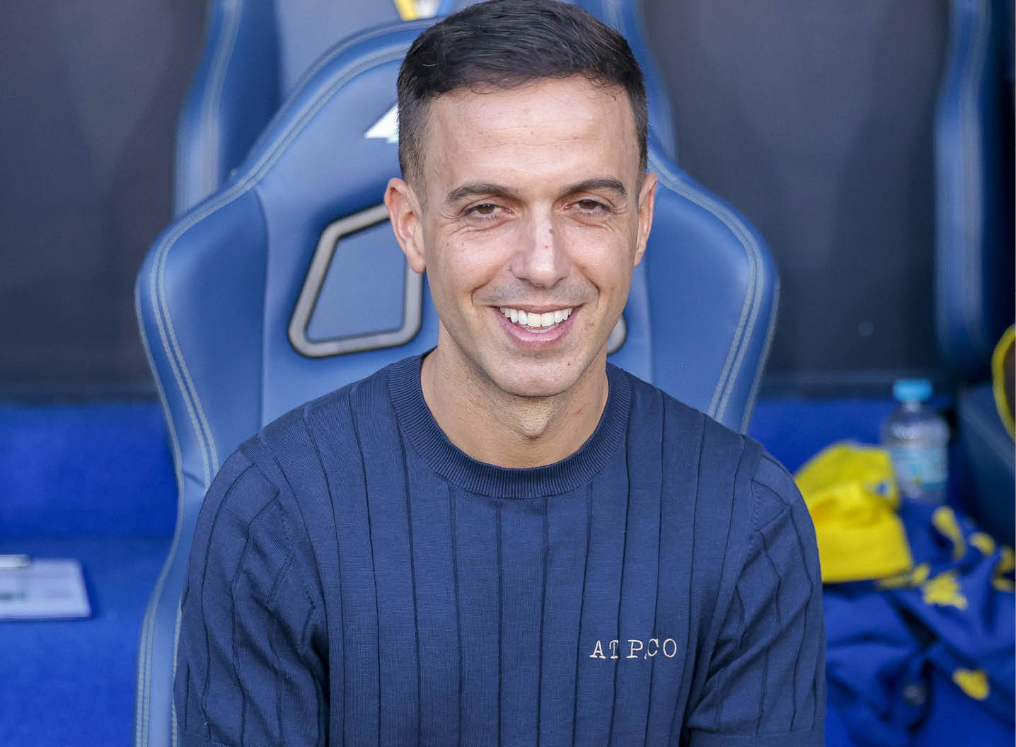 A person wearing a dark blue striped shirt with the text “AT.P.CO" is sitting and smiling in a sports stadium with another person partially visible in sportswear in the background. Descripci n generada con IA