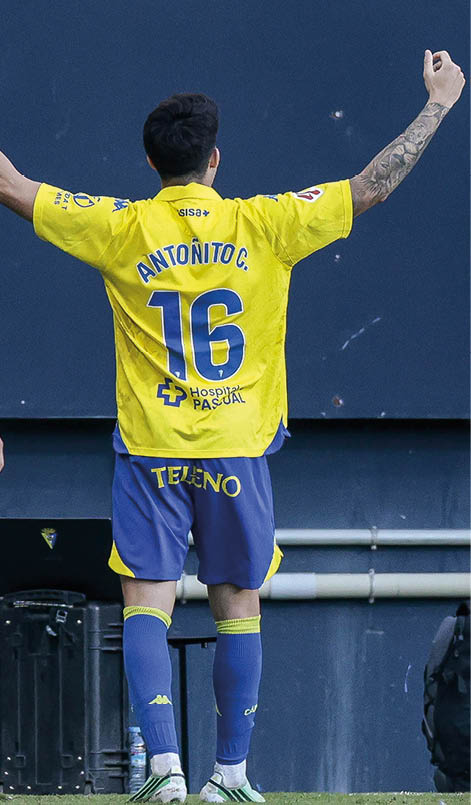 A person wearing a yellow and blue sports uniform with the name “ANTONITO C." and number 16 on the back is raising both arms while standing on a sports field. Descripci n generada con IA