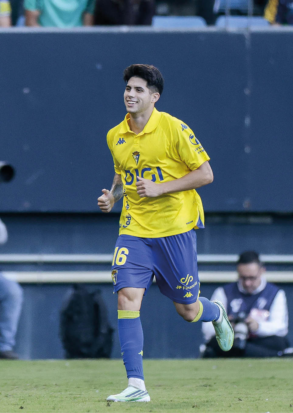 A person wearing a yellow sports jersey and blue shorts is running on a soccer field with a smile. Descripci n generada con IA