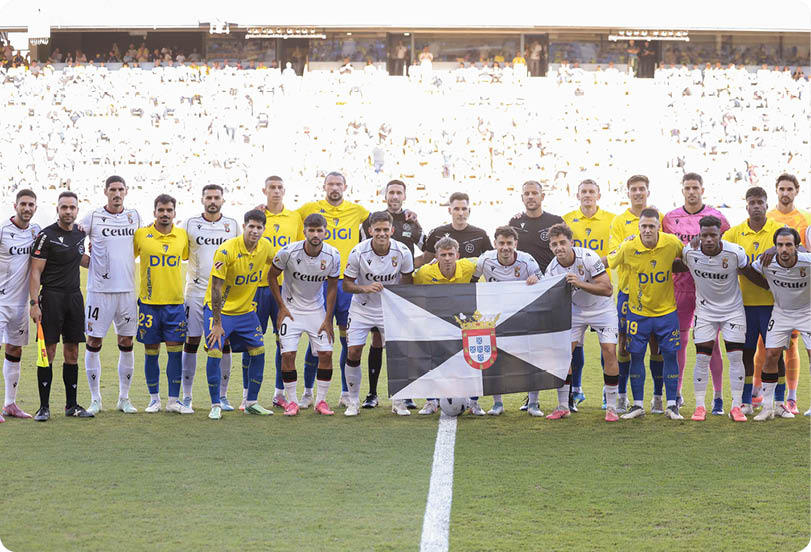Two soccer teams wearing yellow and white uniforms stand together on a soccer field holding a black and white flag with a crest in the center, with a large crowd in the background. Descripci n generada con IA