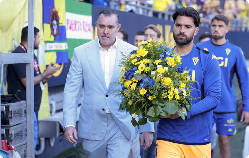 A person in a light blue suit walks beside a person in a blue sports jersey and yellow shorts holding a large bouquet of yellow and blue flowers, with other people in similar sports attire in the background. Descripci n generada con IA